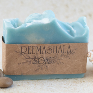 Blue, ocean breeze-scented soap bar flanked by seashells and pebbles.